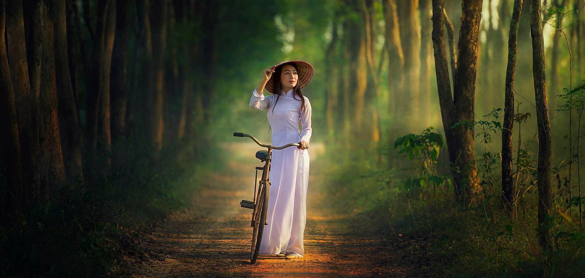 Ung vietnamesiske kvinde i traditionel tøj med cykel på sti kigger på træer i skoven