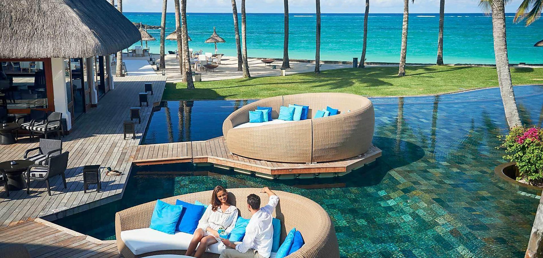 Ung par sidder og snakker i loungesofa, som er bygget på bro i svimmingpool med palmetræer og Mauritius strand i baggrund på resortet Constance Belle Mare Plage 